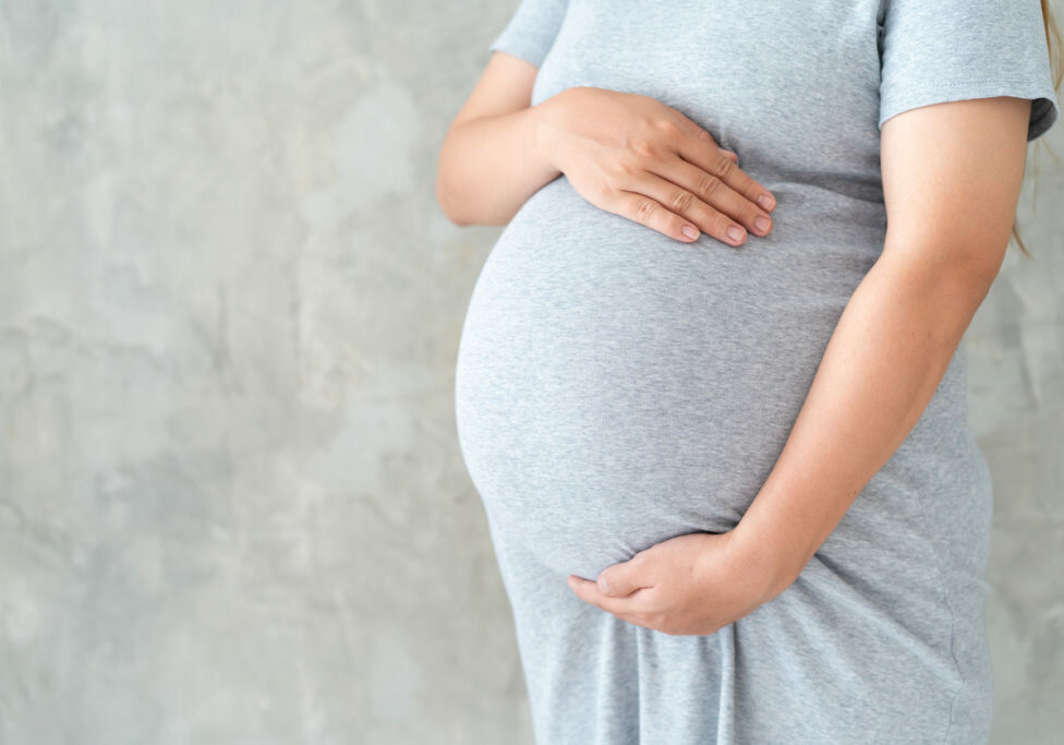 Asian pregnant woman in grey pregnant dress holds hands on her big belly close up on loft concrete wall background.  Concept of healthy pregnant woman, prenant woman's portrait on concrete wall with copy space.