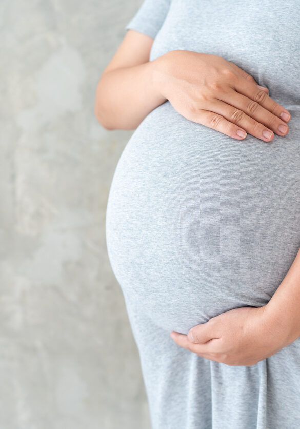 Asian pregnant woman in grey pregnant dress holds hands on her big belly close up on loft concrete wall background.  Concept of healthy pregnant woman, prenant woman's portrait on concrete wall with copy space.